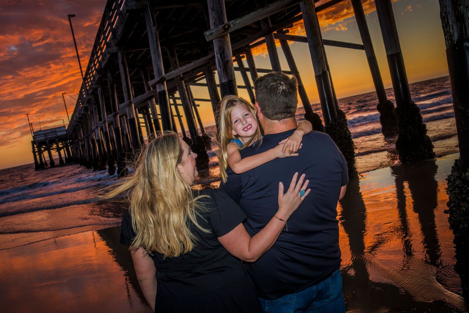 A couple embraces under a wooden pier at sunset with dramatic orange clouds reflecting on the ocean waves.