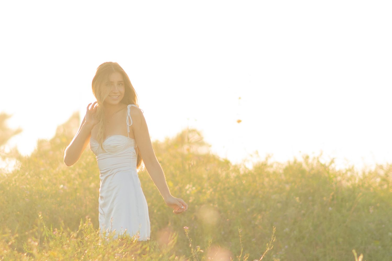 A smiling young woman in a white dress stands in a sunlit field, bathed in warm golden light.
