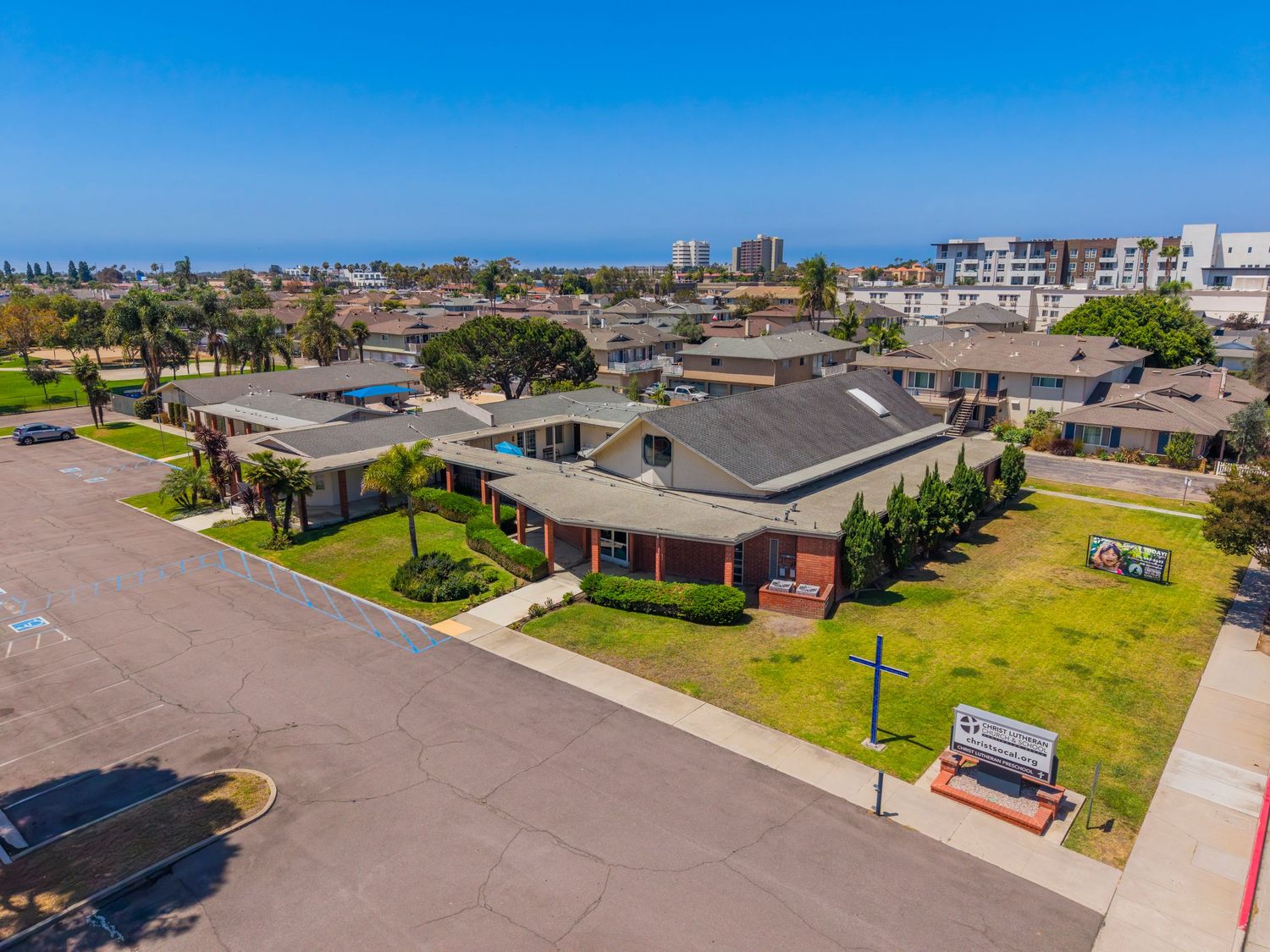 Aerial view of a low-rise commercial building with green lawn, parking lot, and ocean visible in the background under blue sky.