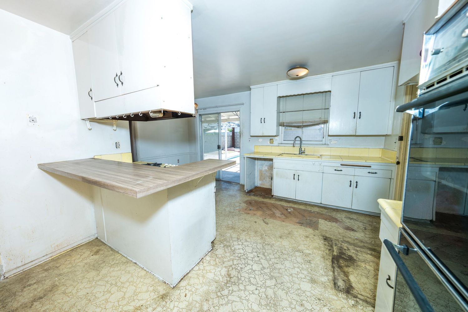 Outdated kitchen with white cabinets, wooden peninsula counter, tile flooring, and stainless steel oven needing renovation.