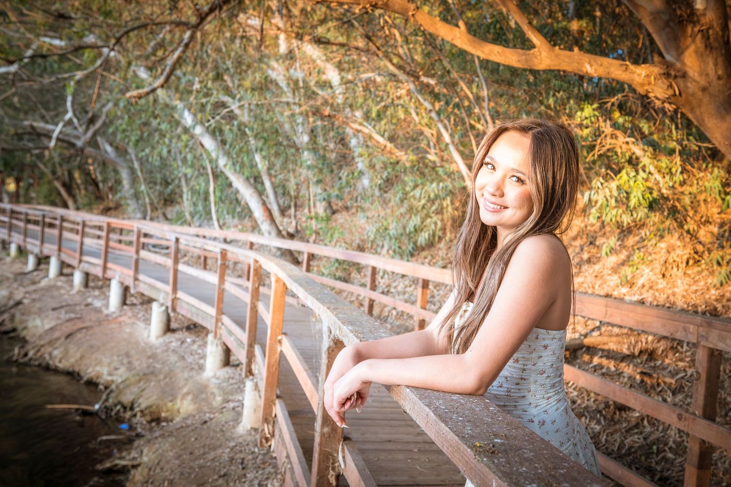 Young woman smiling on a wooden bridge surrounded by autumn trees in warm golden light.
