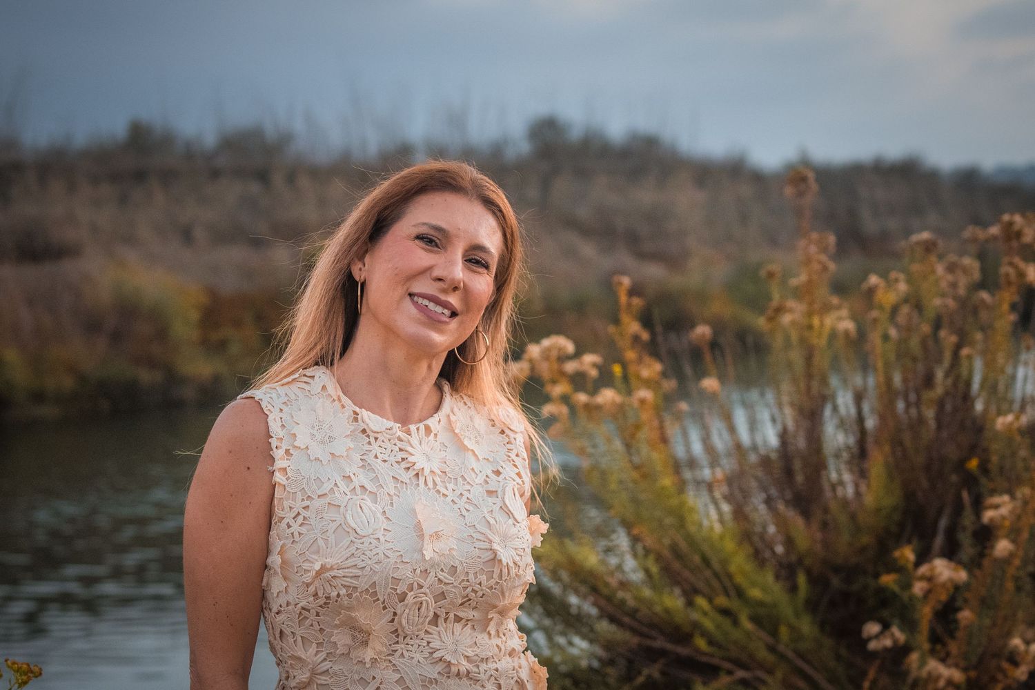 A bride in a white lace wedding dress poses outdoors during golden hour near a lake.