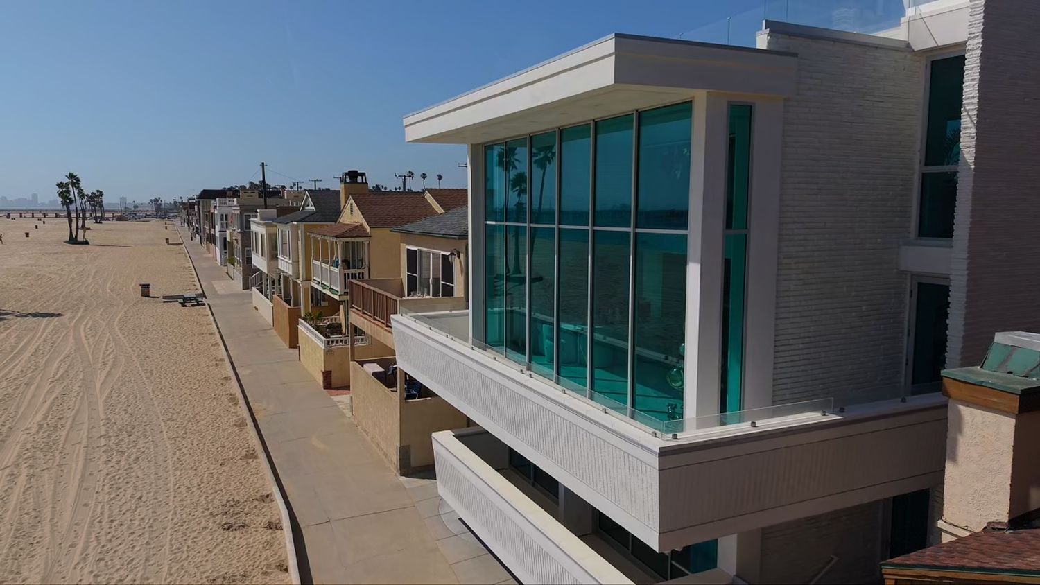 Modern beachfront home with floor-to-ceiling glass windows overlooking a sandy beach boardwalk.