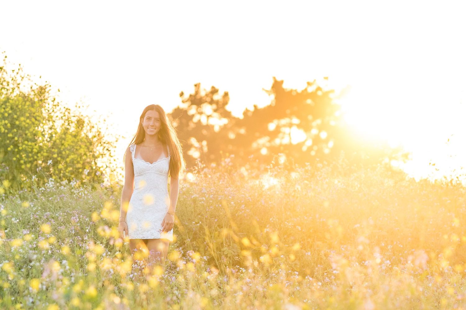 Young woman in white dress standing in golden sunlit meadow with wildflowers and glowing backlight at sunset.