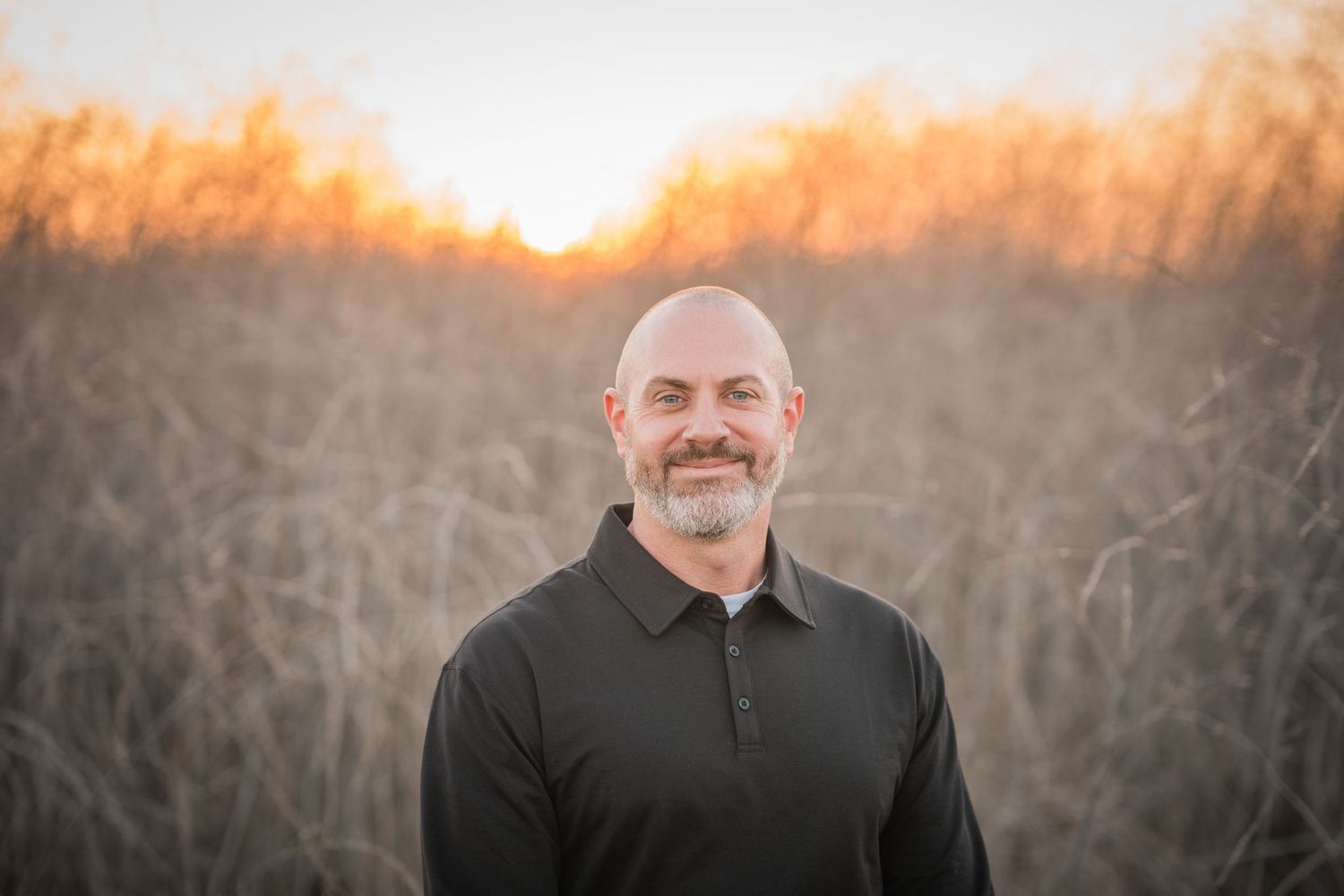 Person in black jacket poses outdoors during golden sunset with bare trees in background.