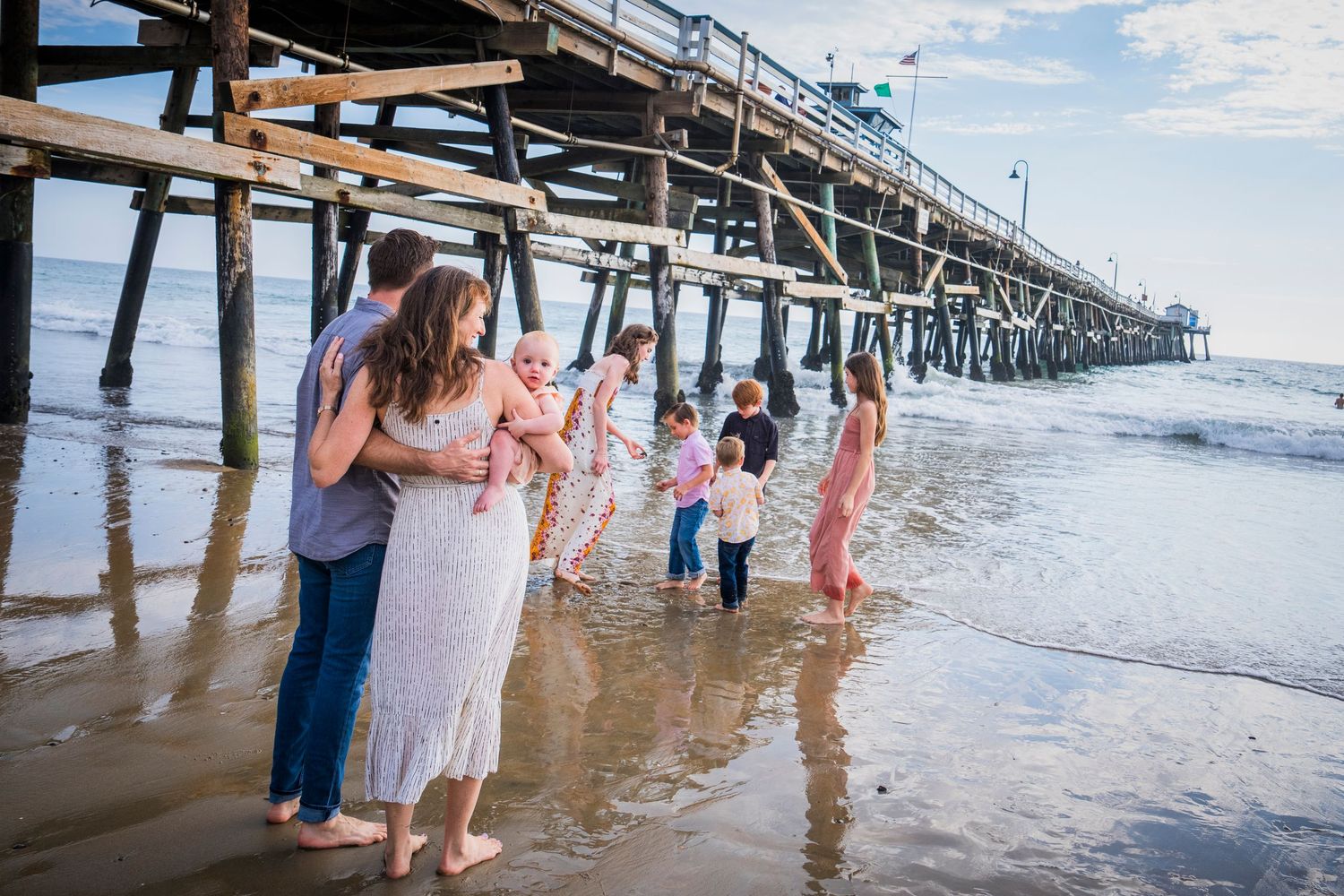 People stroll along a wooden pier extending into the ocean as waves crash against the pilings on a sunny day.