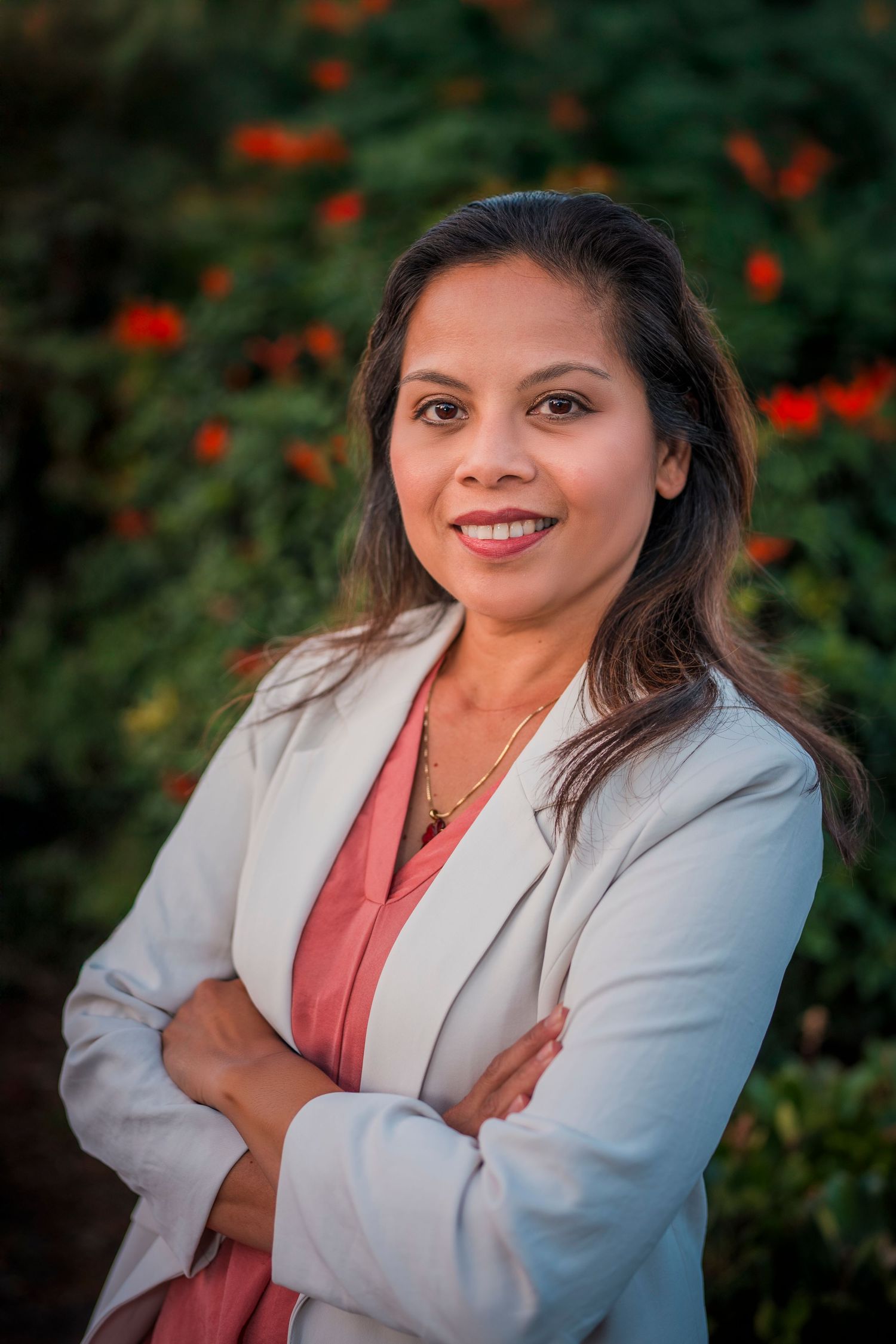 A woman in a grey blazer and coral blouse poses outdoors against a background of red flowers and greenery.