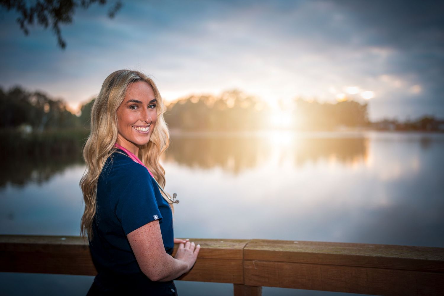 Smiling blonde woman in navy scrubs poses by a sunlit lake with golden hour reflections at sunset.