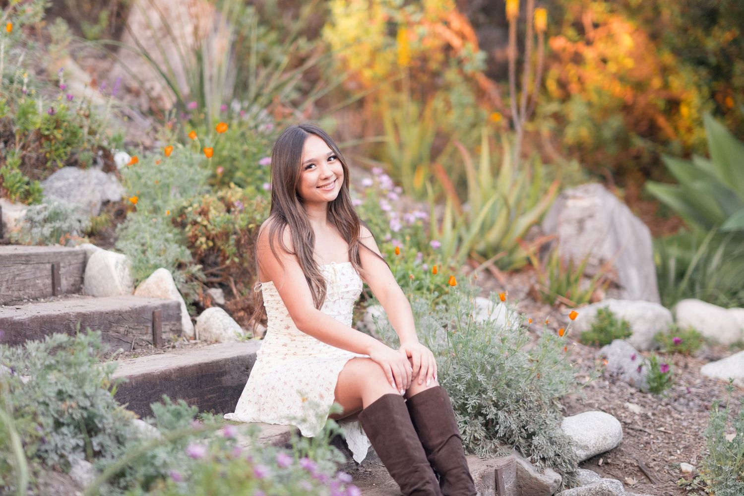 Young woman in white dress and brown knee-high boots sitting in a colorful garden with rocks and autumn foliage.