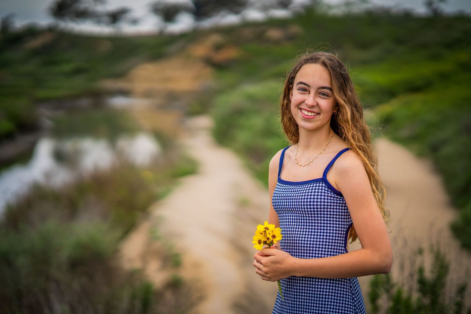 Graduate holding yellow flowers on dirt path with green hills in background.