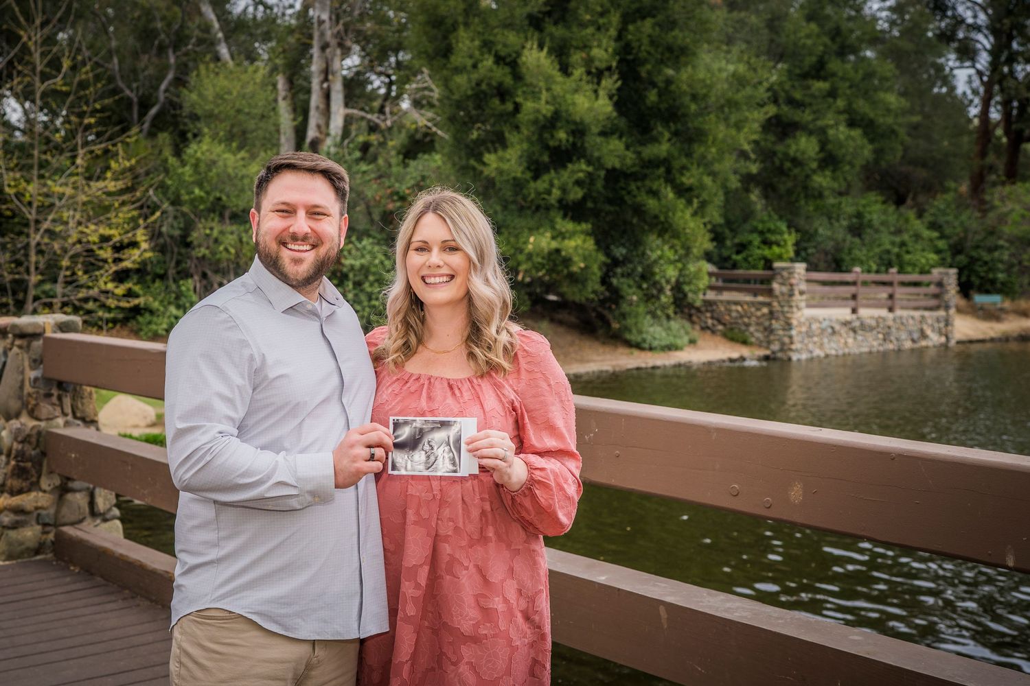 Couple announcing child while standing on wooden bridge over pond.