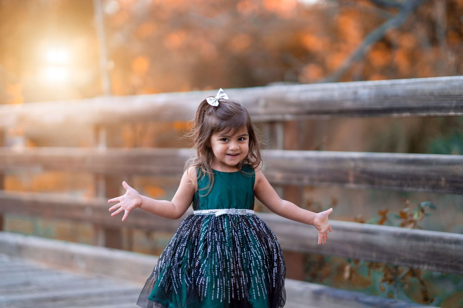 Child in emerald green dress twirling on wooden bridge during autumn sunset.
