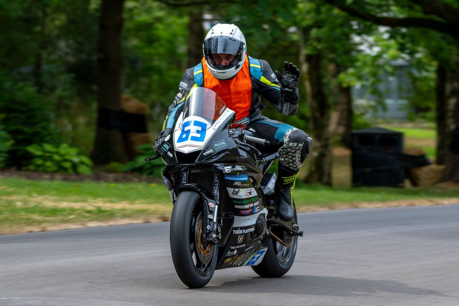 Motorcycle racer number 63 wearing orange vest navigating a tree-lined road racing circuit corner.