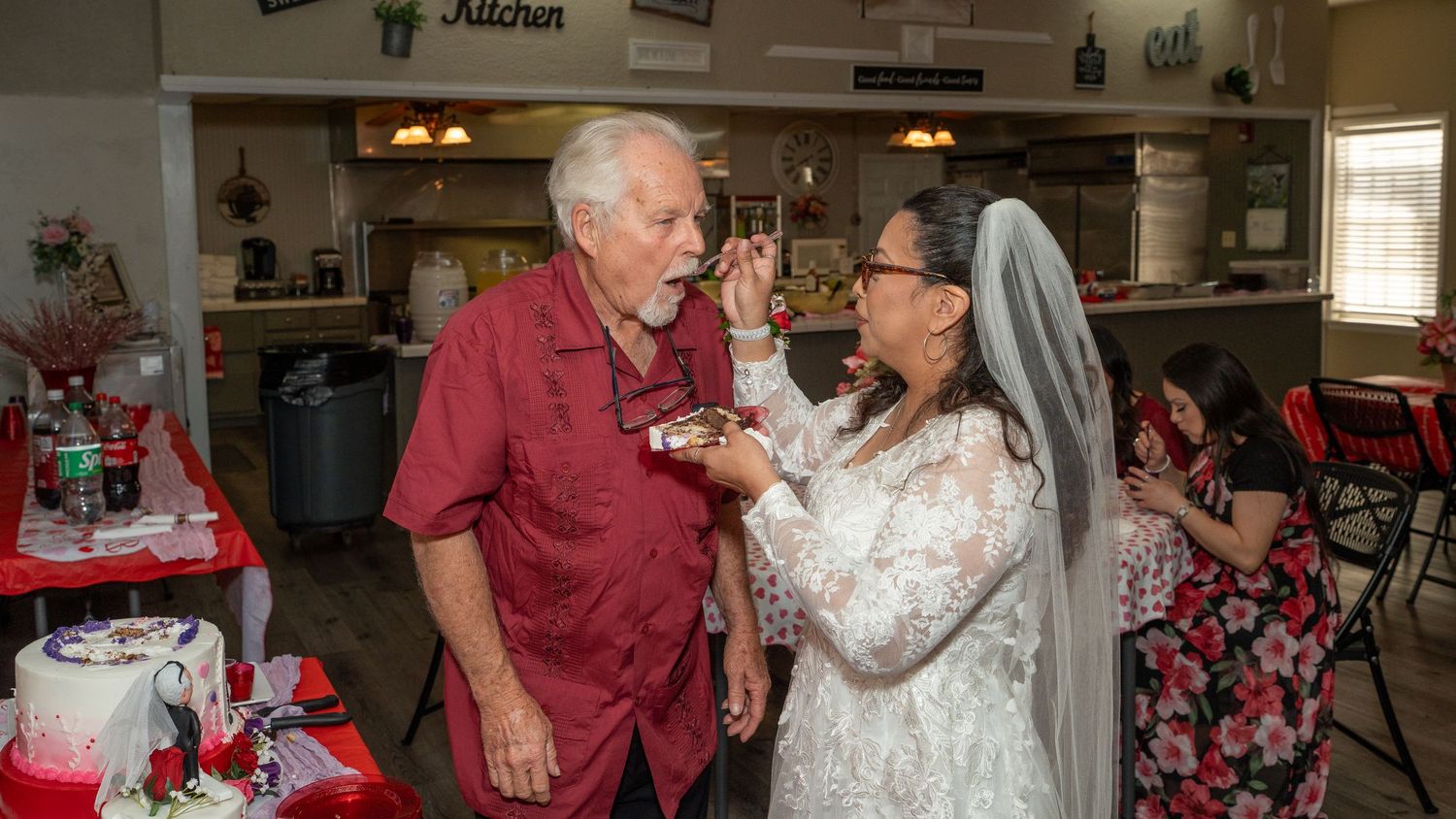 Elderly couple sharing wedding cake at their reception, bride in white lace gown and veil, groom in red shirt.