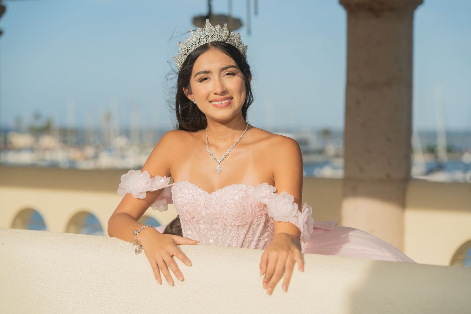 Young Latina woman in pink quinceañera gown and tiara poses gracefully at a waterfront venue on a sunny day.