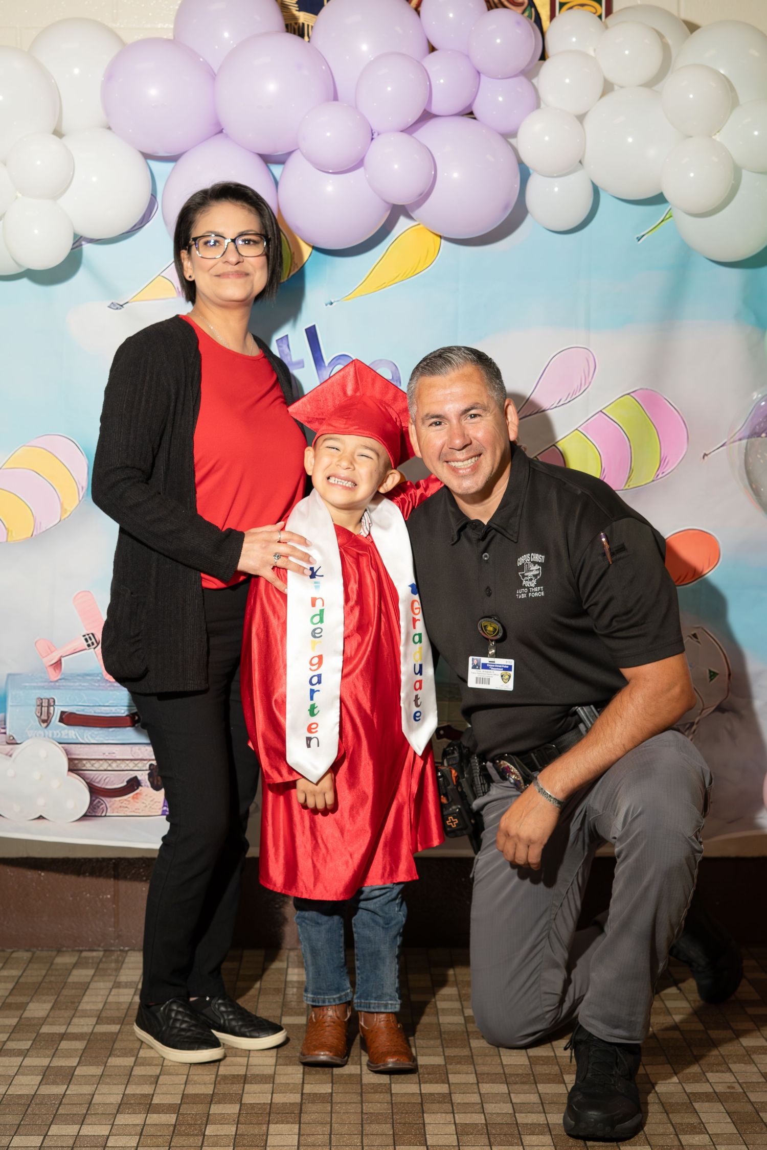 A young child in red graduation cap and gown poses with two proud parents in front of a colorful balloon backdrop.