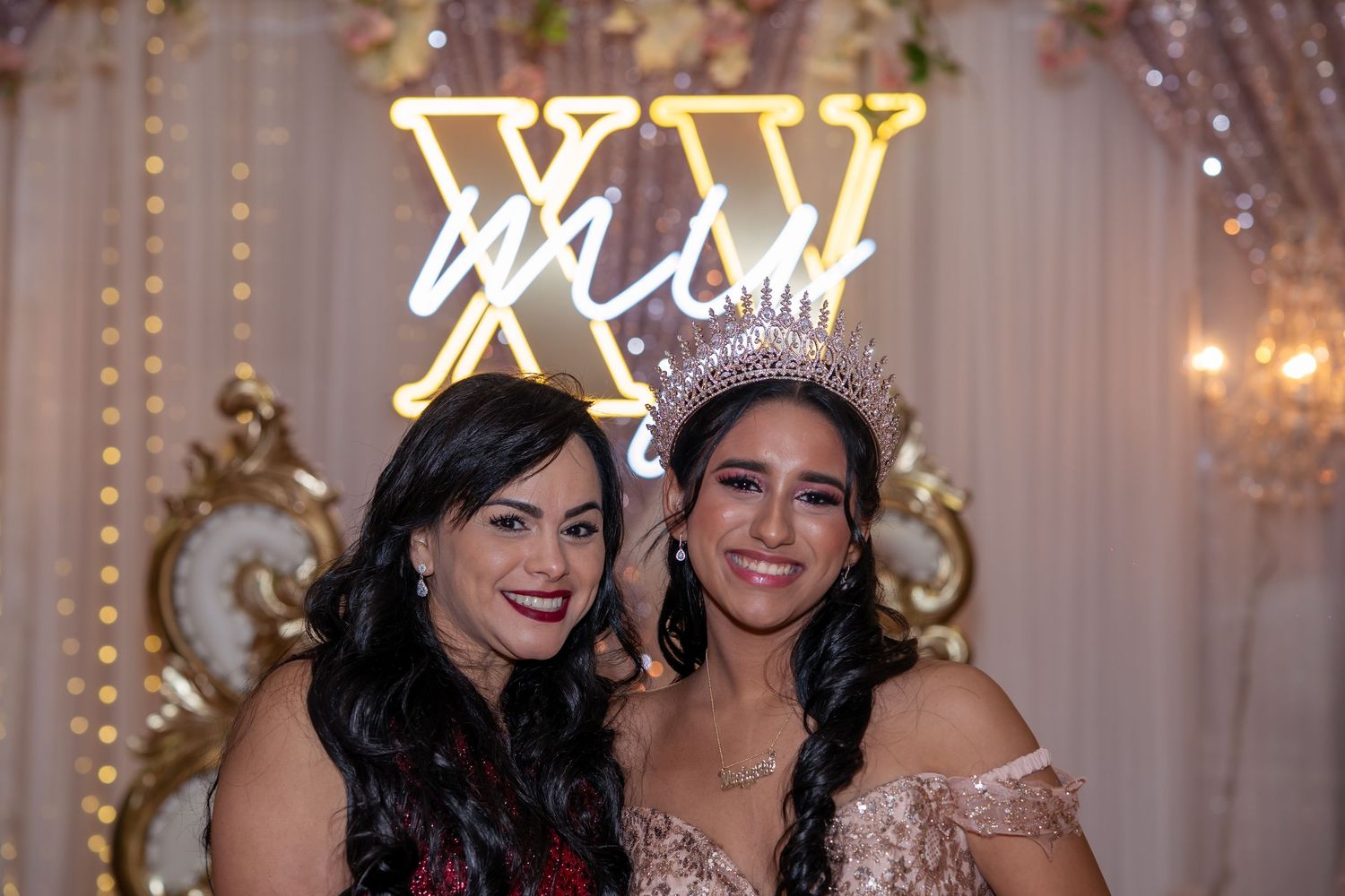 Two women pose together at a quinceañera celebration, one wearing a crown and gown, in front of an XV neon sign.
