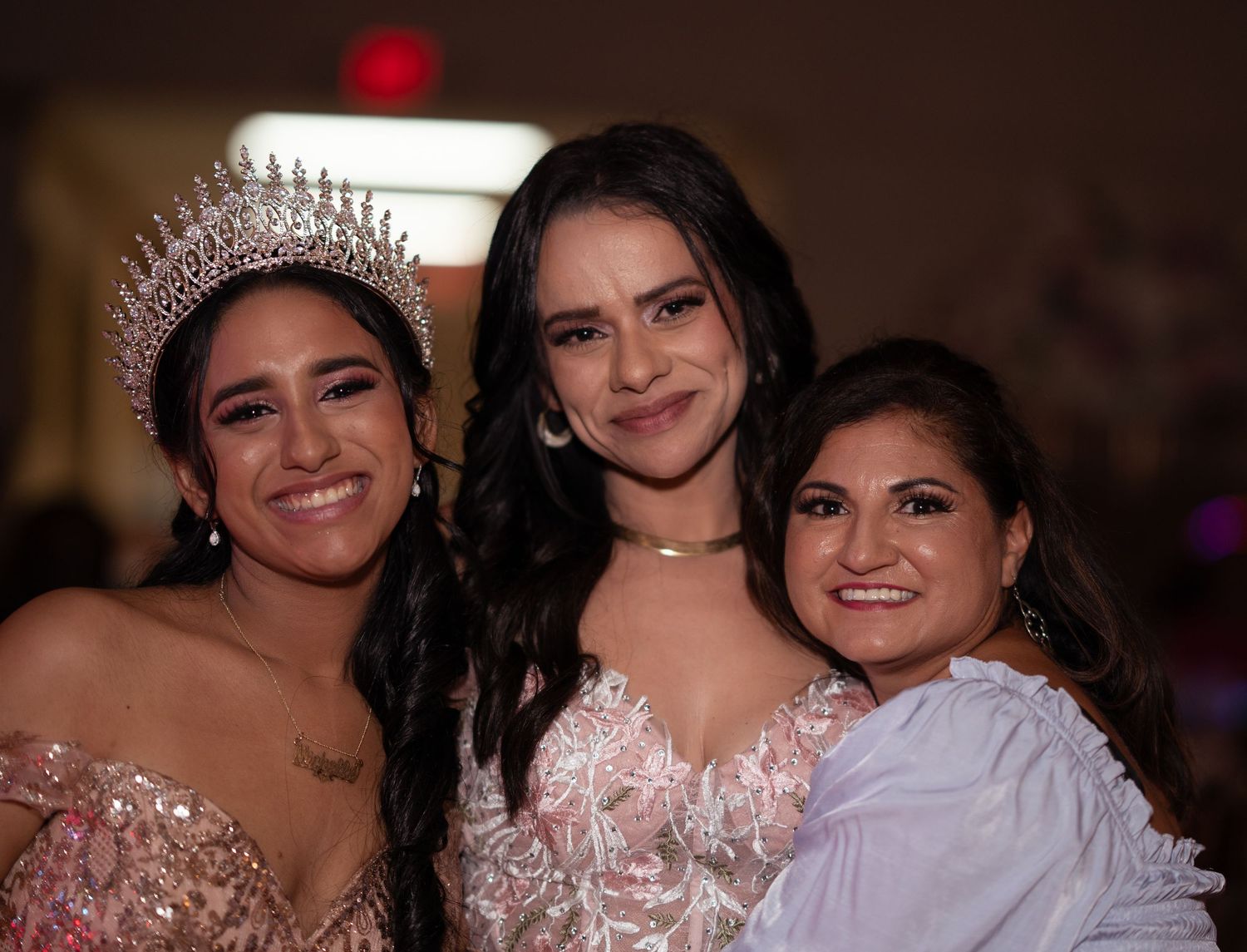 Three smiling women pose together at a quinceañera, with the honoree wearing a sparkling tiara and pink gown.