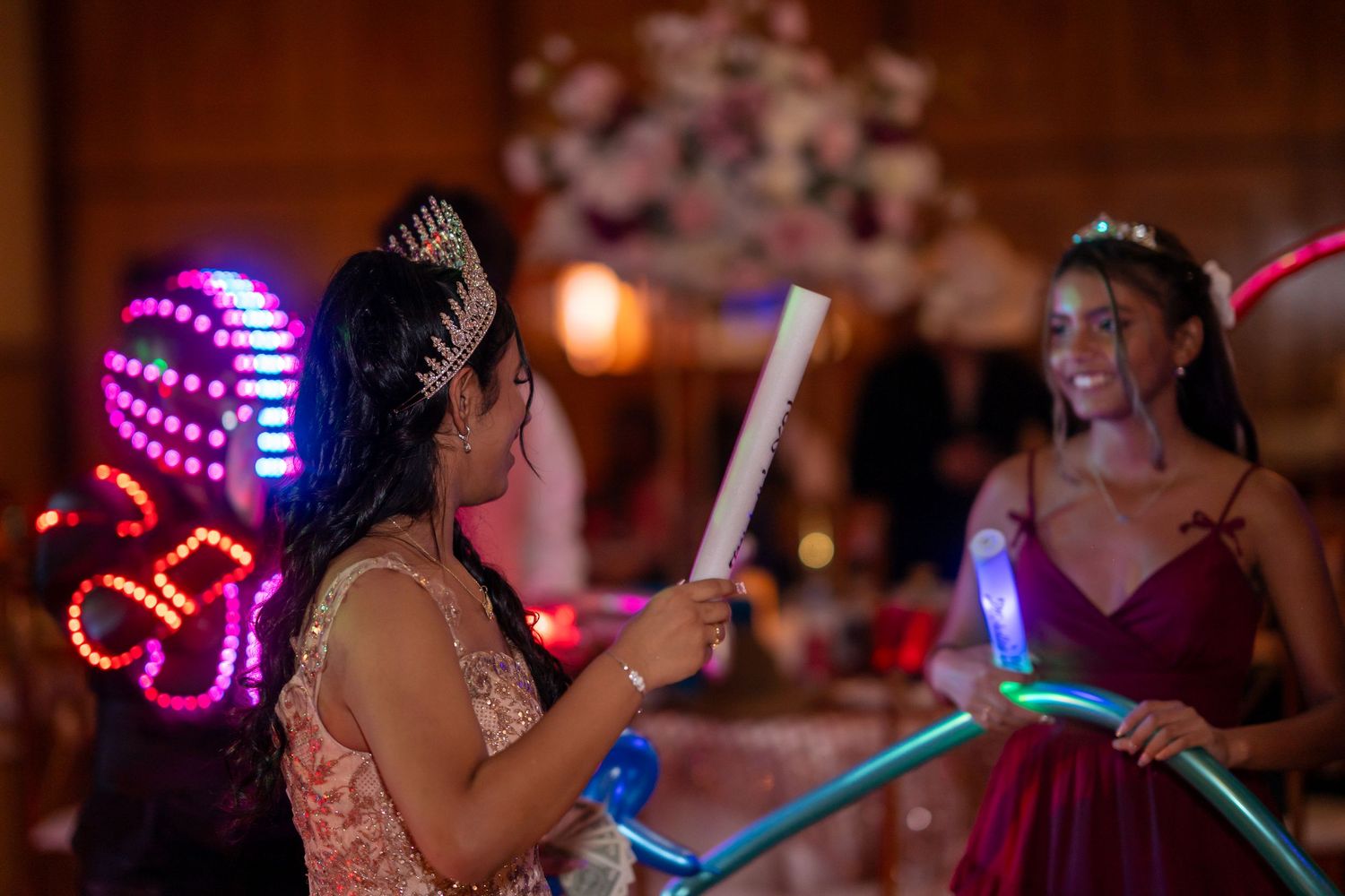 Two girls in formal dresses celebrate a quinceañera with glowing light-up accessories and neon decorations.