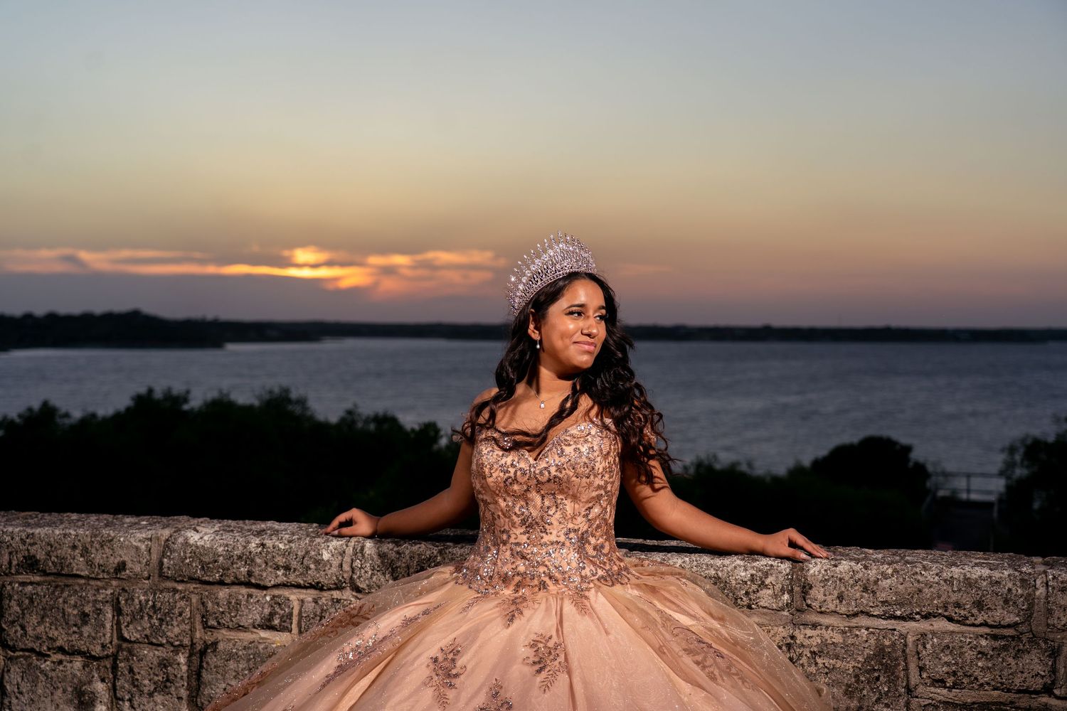 A person in a blush pink formal gown and beanie poses at sunset by a stone wall overlooking a lake.