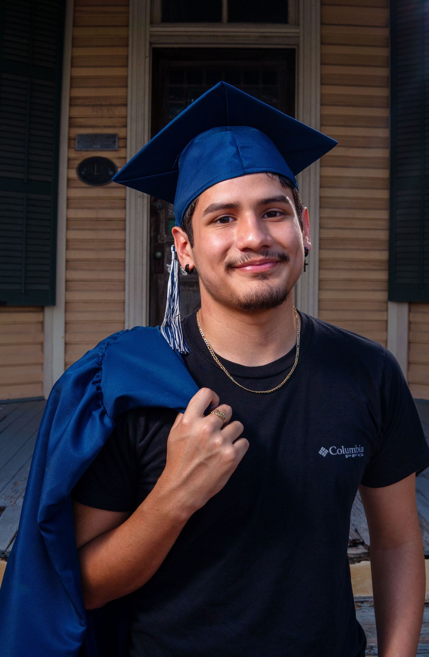 A graduate poses in front of a house wearing a blue cap and gown with a black t-shirt underneath.