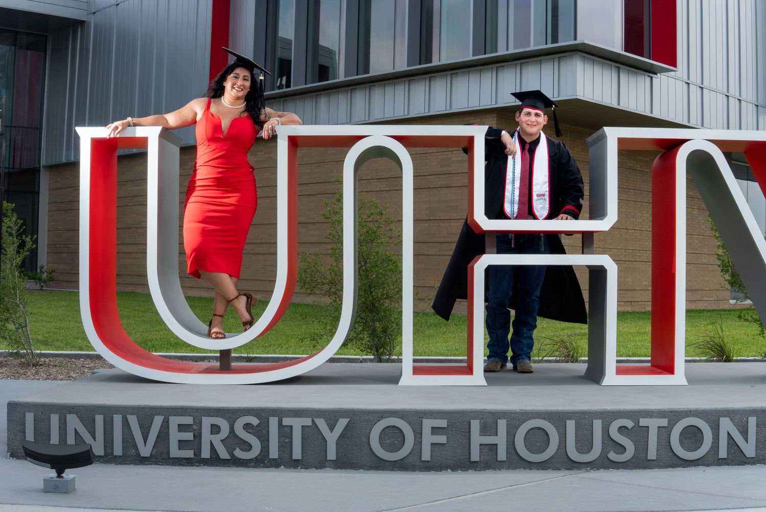 Two graduates pose creatively on the UH letters at the University of Houston entrance sign for a graduation photo.