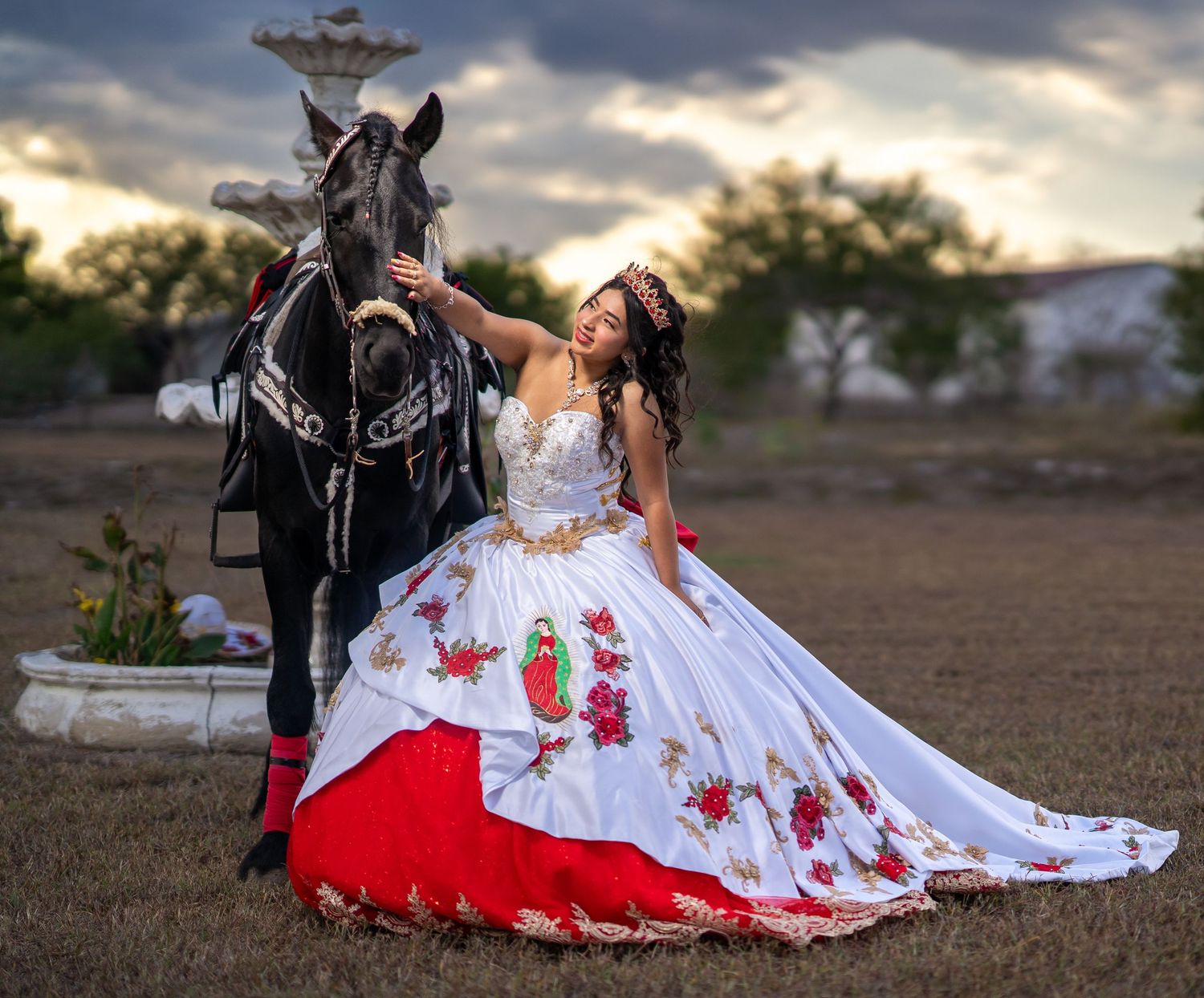An elegant white and red floral gown contrasts beautifully with a black horse against a dramatic sunset sky.