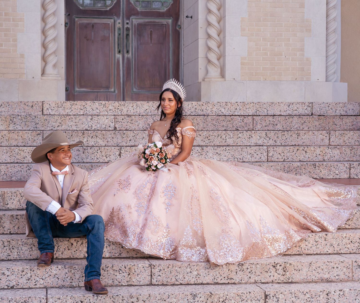 A person in a flowing pink ballgown and cowboy boots sits on stone steps in a romantic outdoor photoshoot.