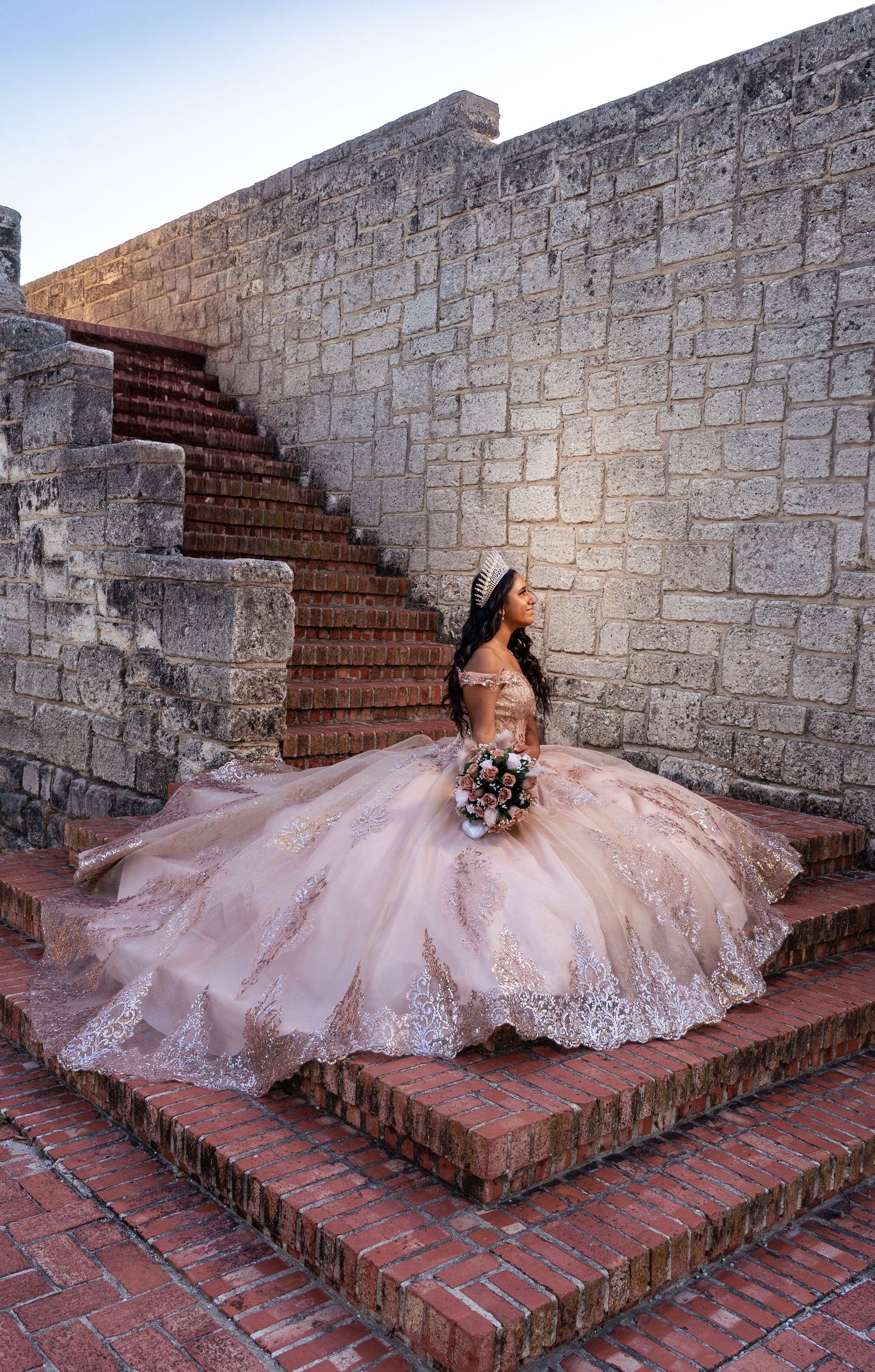 Rose gold quinceañera gown with intricate beading spreads elegantly across brick stairs in a dramatic pose.