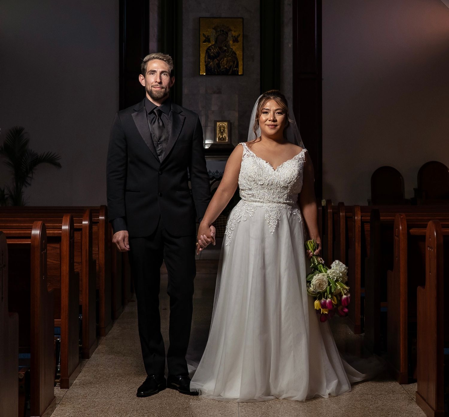 Bride and groom holding hands in church aisle, bride in white lace gown with bouquet, groom in black suit.