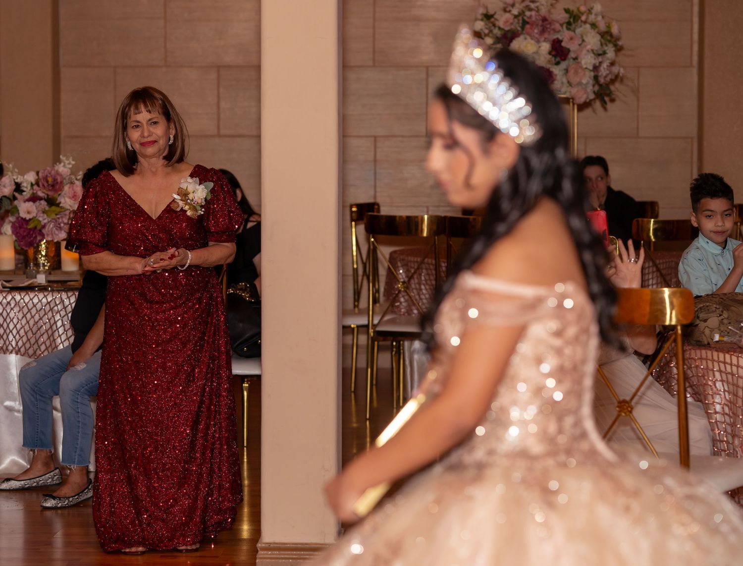 A woman in a red sequin gown watches a quinceañera girl in a gold gown during a celebration.