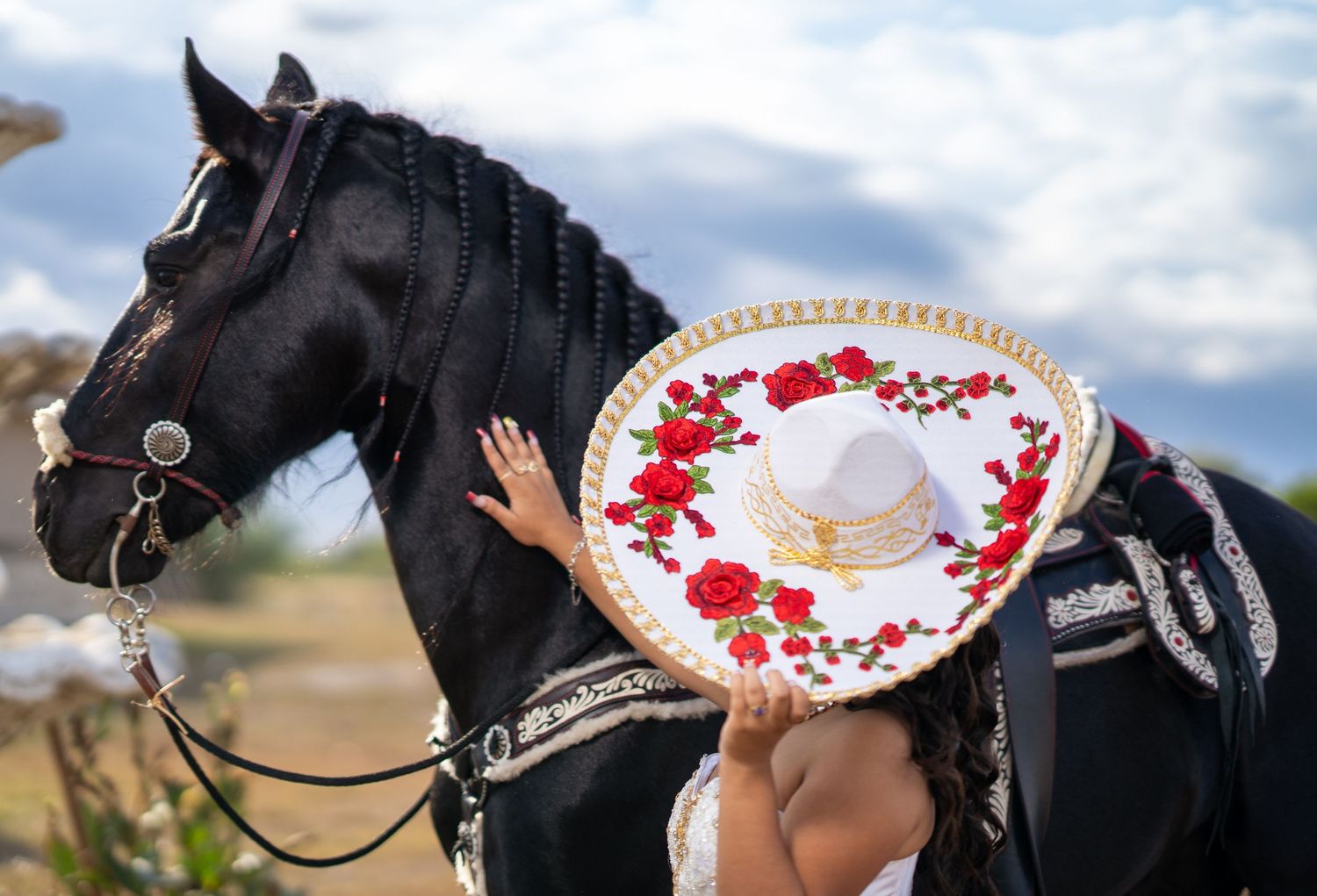 A black horse stands next to someone wearing a white dress with red floral accents in an outdoor setting.