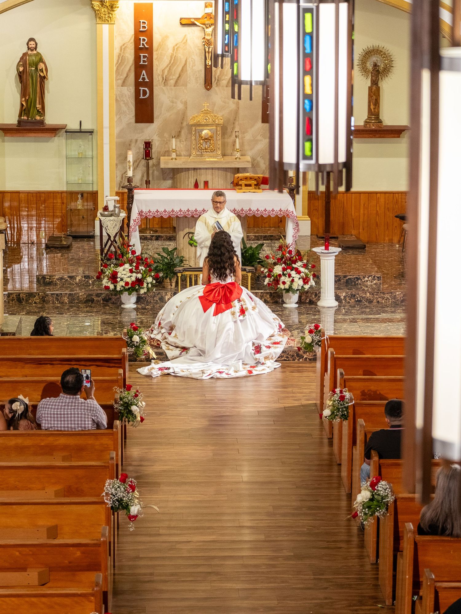 A church interior with wooden pews decorated with floral arrangements and a person in white kneeling at the altar.