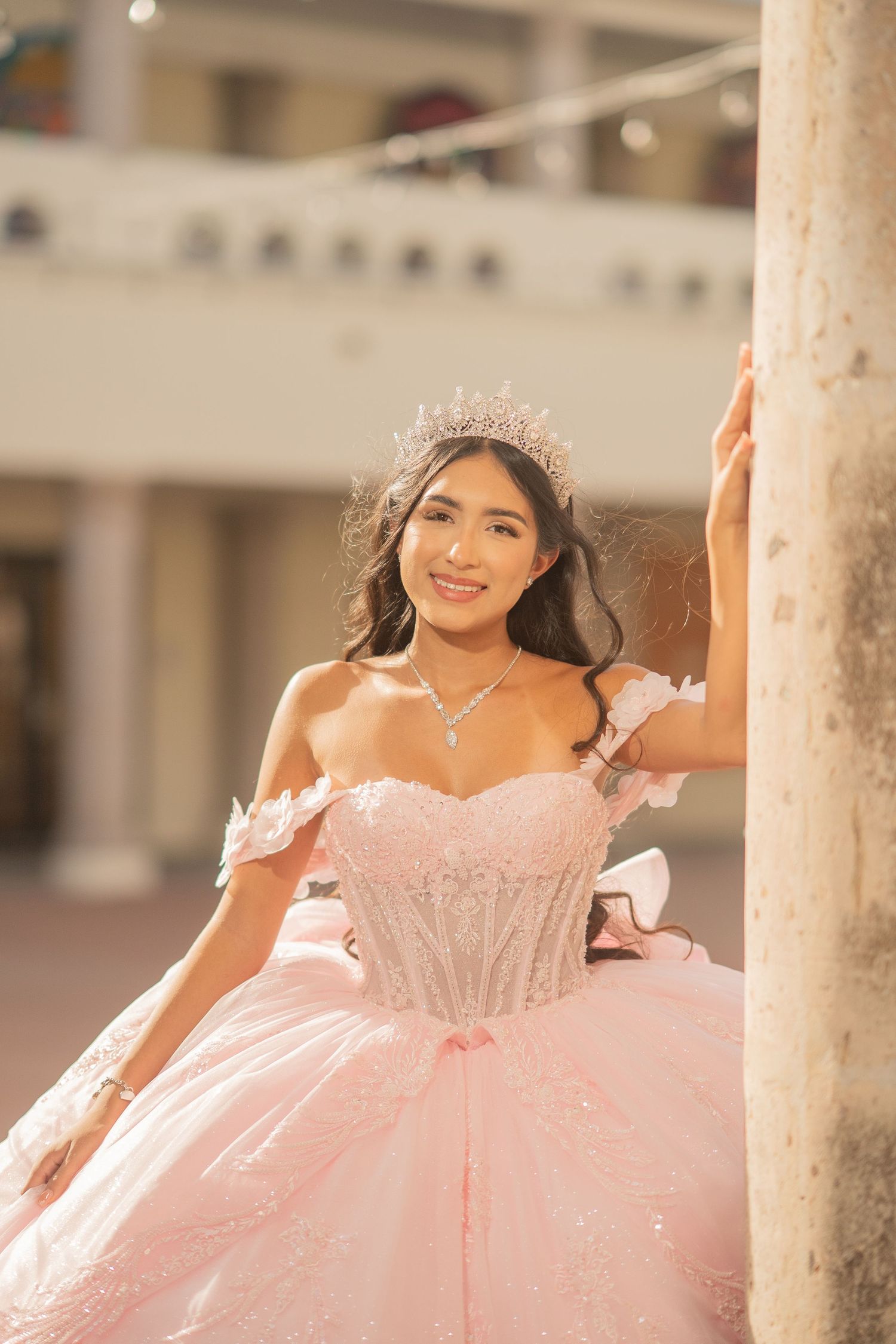 Young Latina woman in pink quinceañera ball gown with crystal crown and necklace posing outdoors.