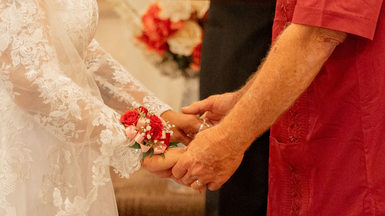 Bride and groom holding hands at wedding ceremony, bride wearing lace dress with floral wrist corsage.