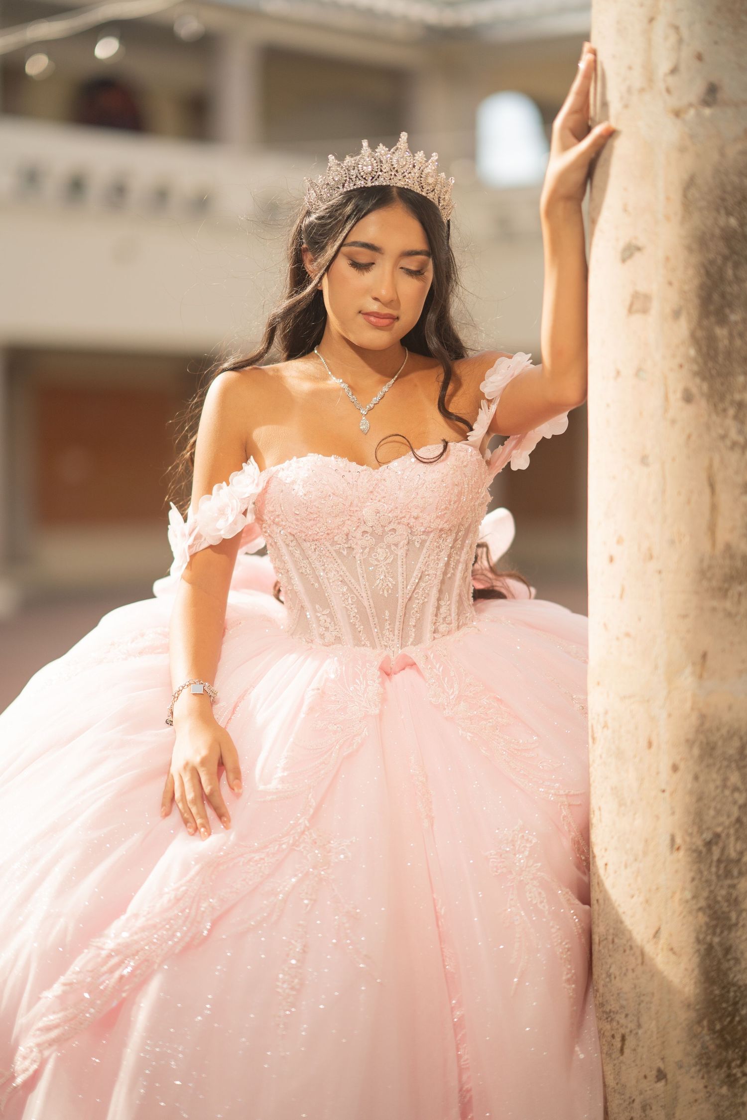 Young woman in pink ball gown and tiara poses gracefully against a column during her quinceañera celebration.