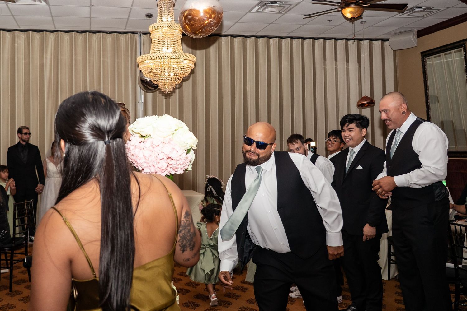 Wedding guests dancing and celebrating at a reception venue with chandelier lighting and floral bouquet.