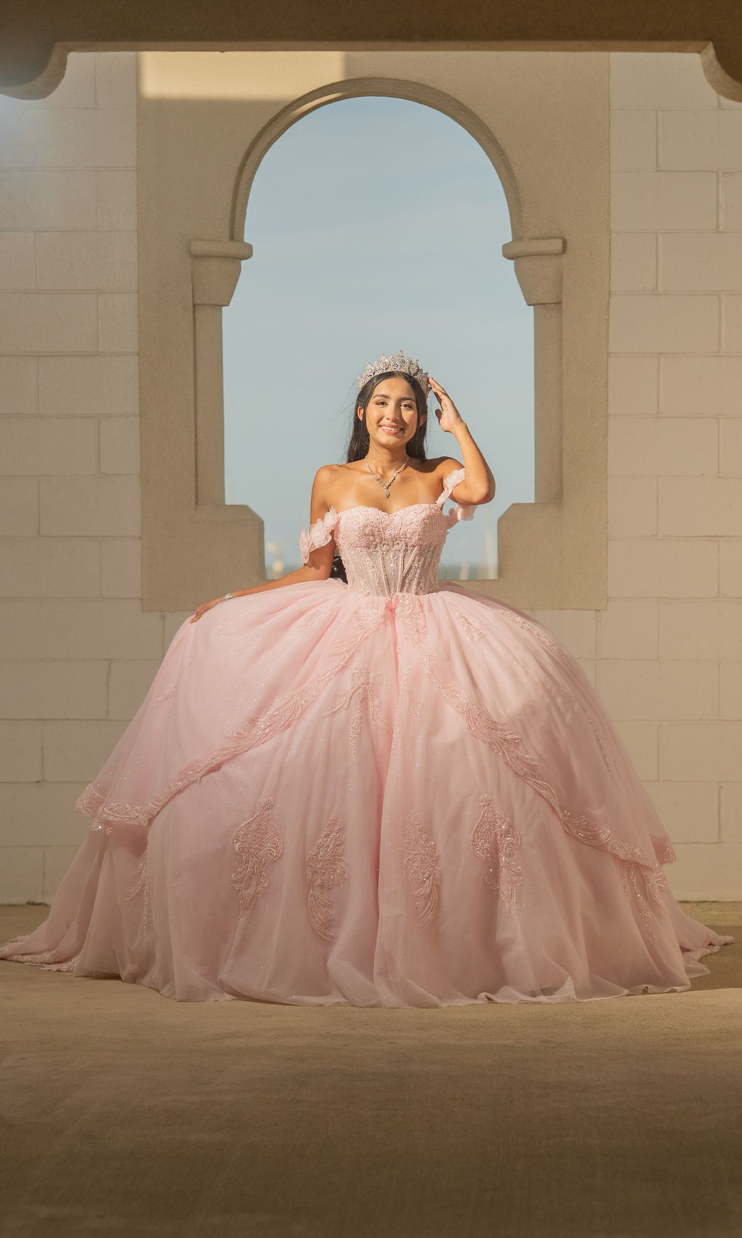 Young woman in pink ball gown quinceañera dress with tiara poses under stone arch.