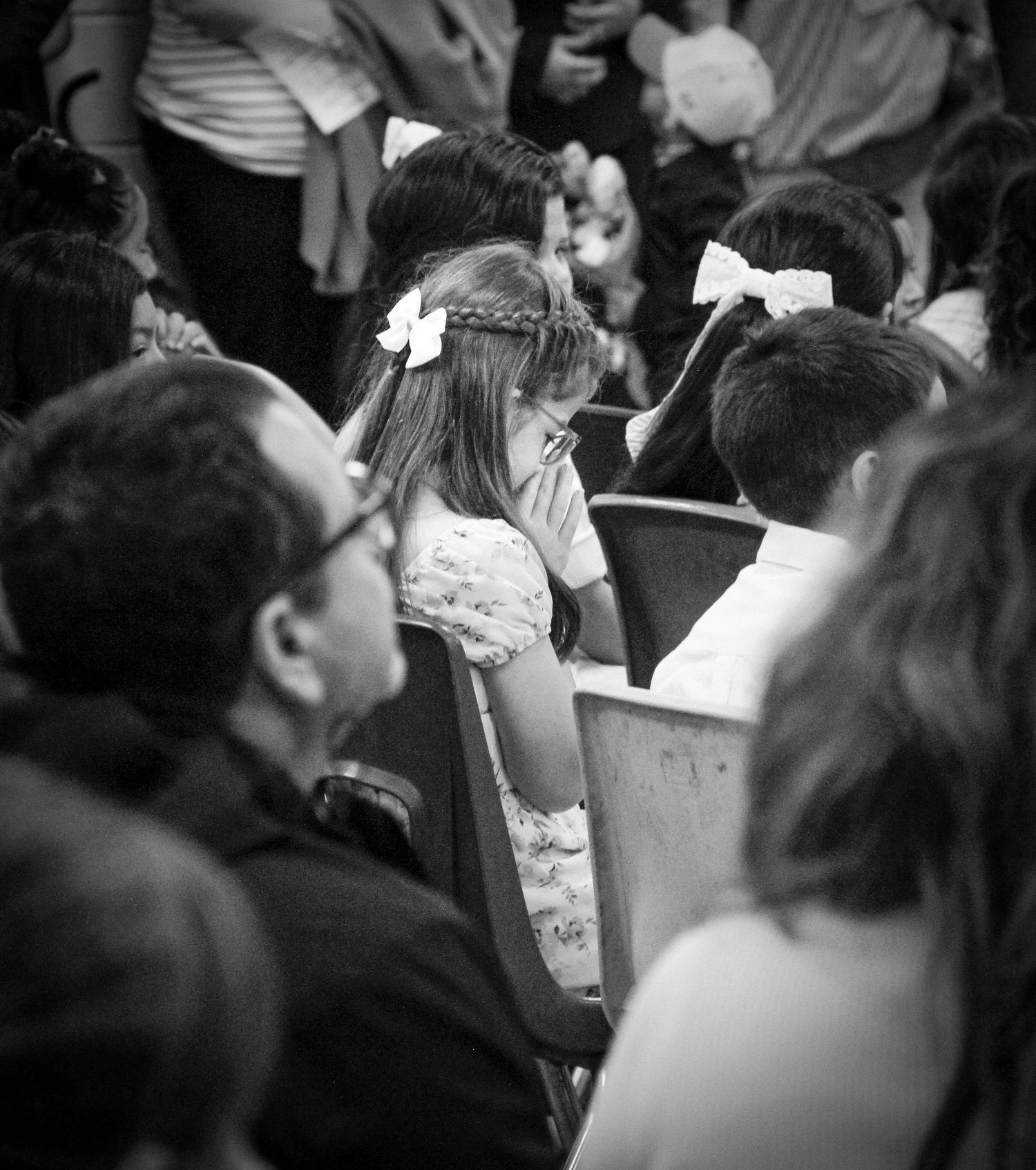 Black and white photo of a young girl with bow praying in church pew among a crowd of people.
