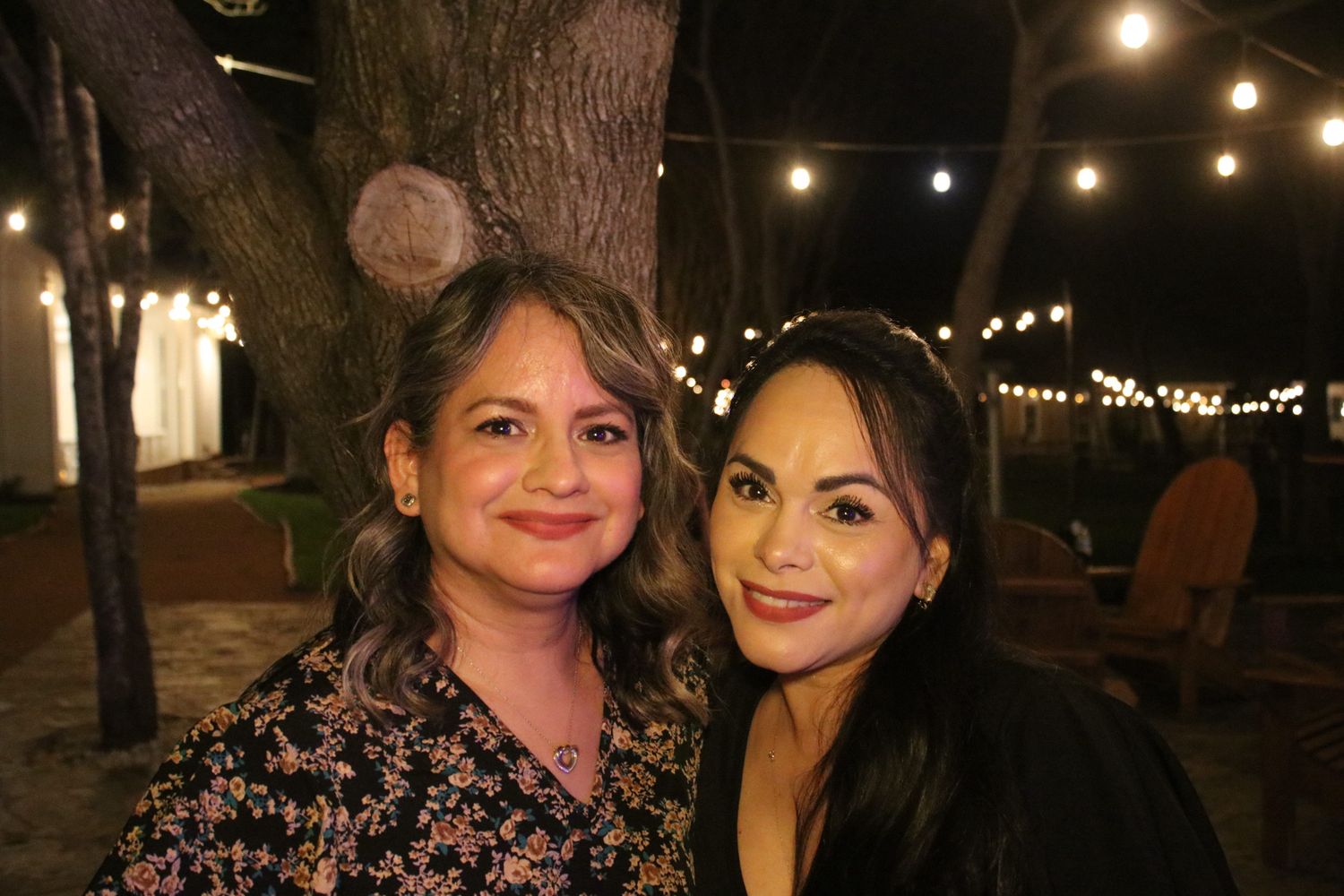 Two women smile at the camera in an outdoor setting with string lights in the background.