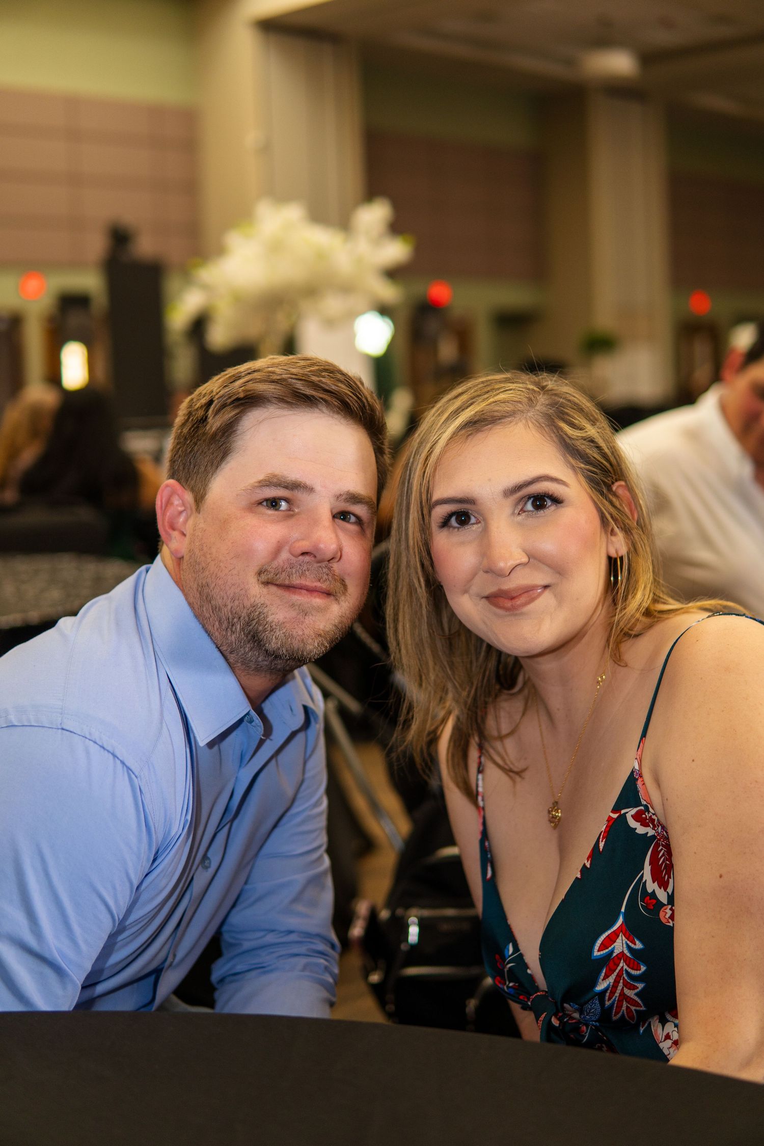 Two people sit together at a table during an indoor event, wearing dressy casual attire in a warmly lit setting.