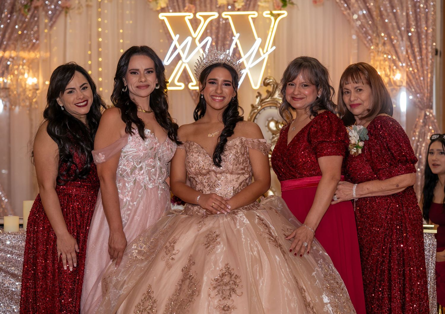 Quinceañera honoree in gold gown and tiara posing with four women in red and floral dresses.