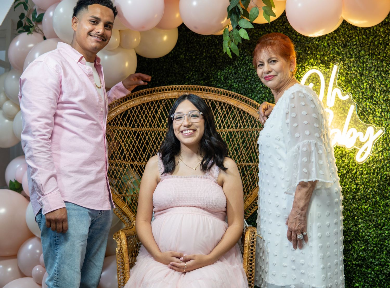 A baby shower celebration with pink and beige balloons, a wicker peacock chair, and a neon 'Oh' sign on a green wall.