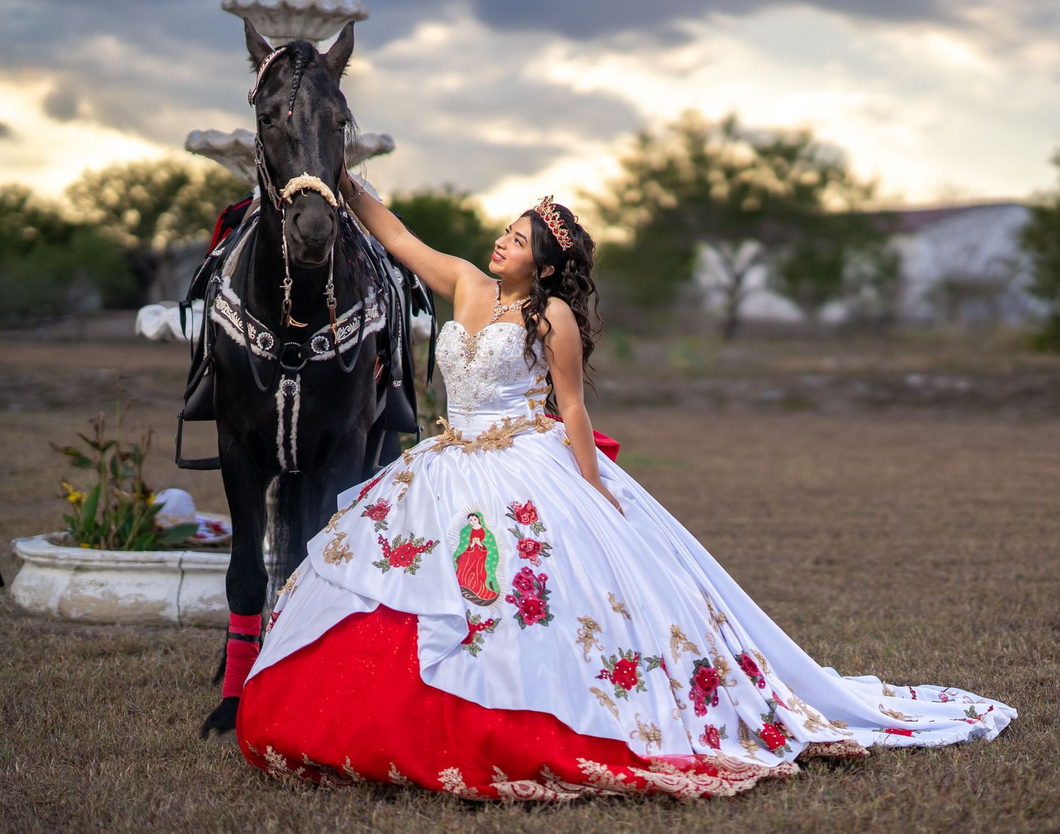 A dramatic western-themed photoshoot featuring a black horse and flowing white dress with red accents against a sunset sky.