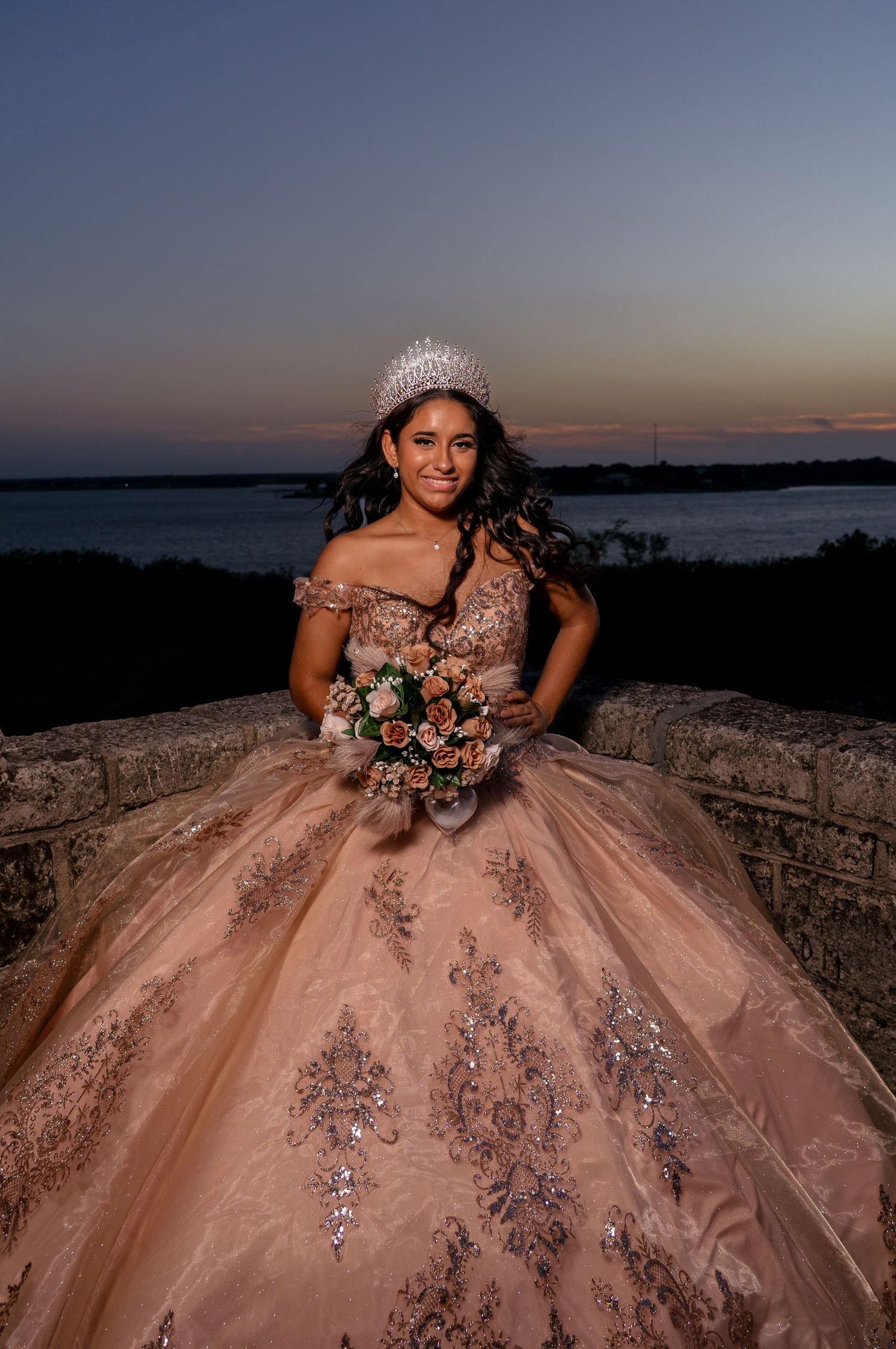 Sparkling champagne ball gown with floral bouquet photographed against sunset waterfront view.