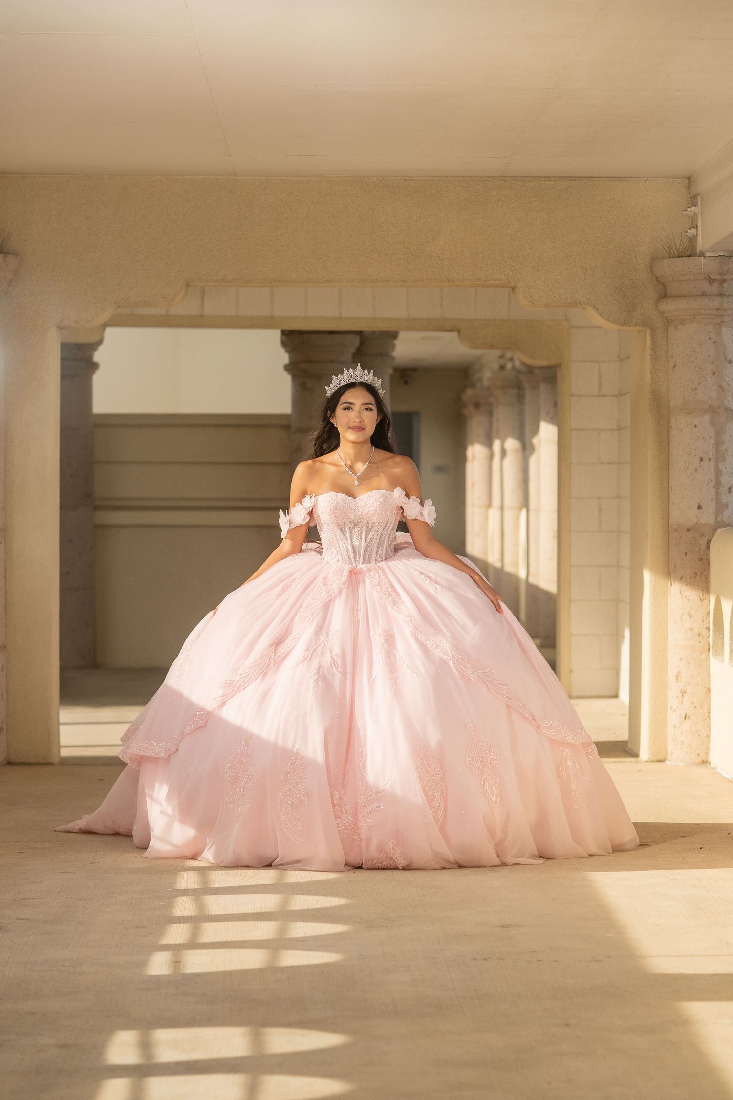 Elegant quinceañera in a blush pink ball gown and tiara stands in a sunlit archway.