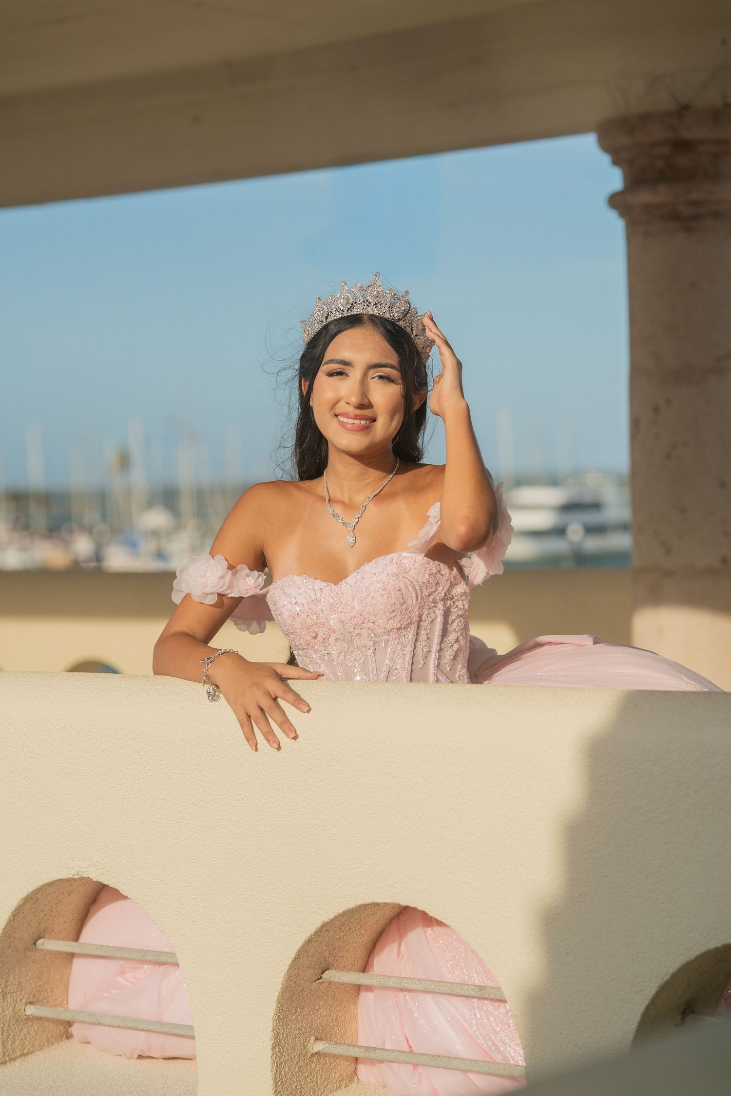 Smiling quinceañera in pink ball gown and crystal tiara leans on arched balcony railing by harbor.