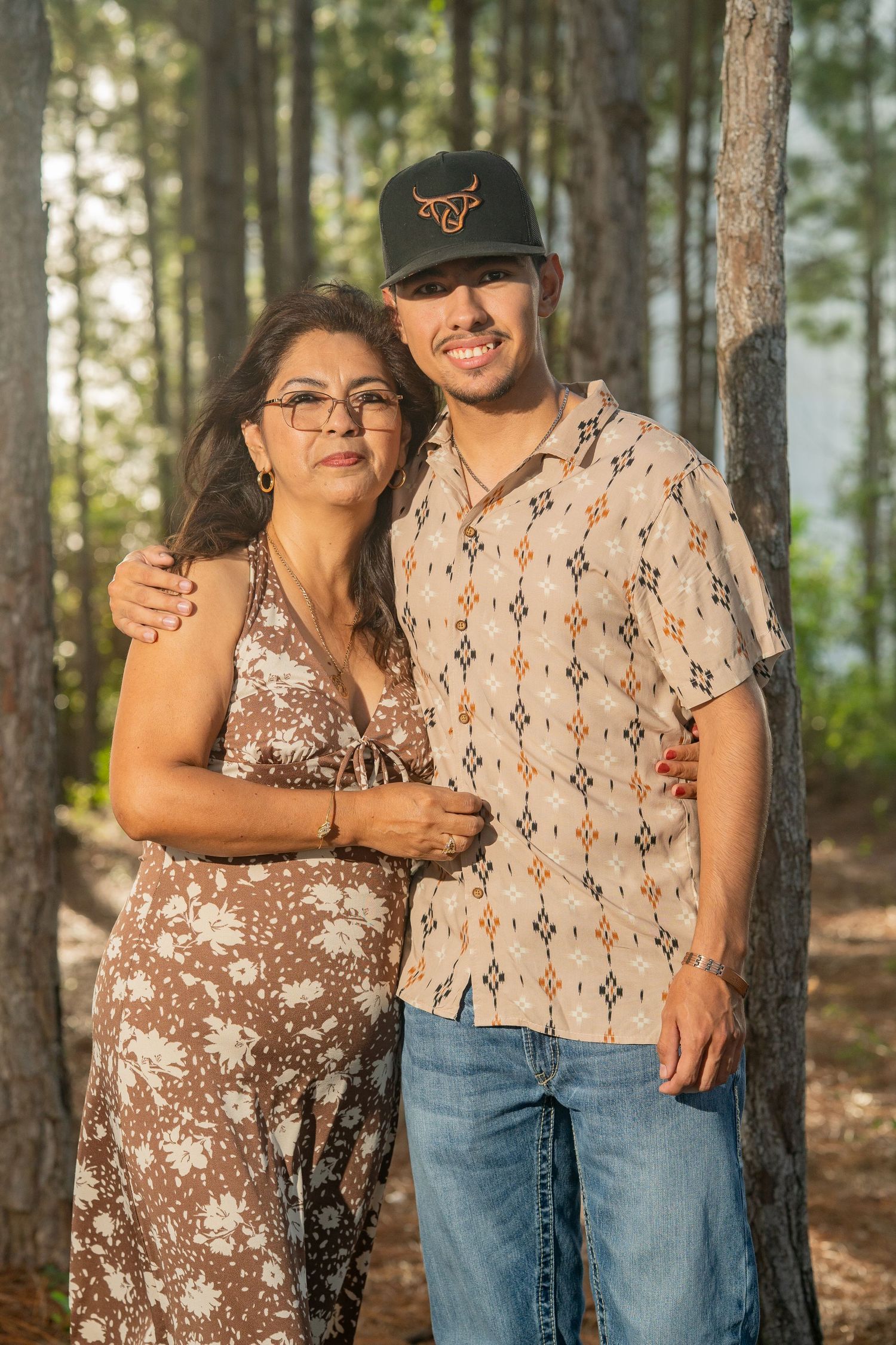 A smiling young man in a patterned shirt and cap poses with an older woman in a floral dress among pine trees.