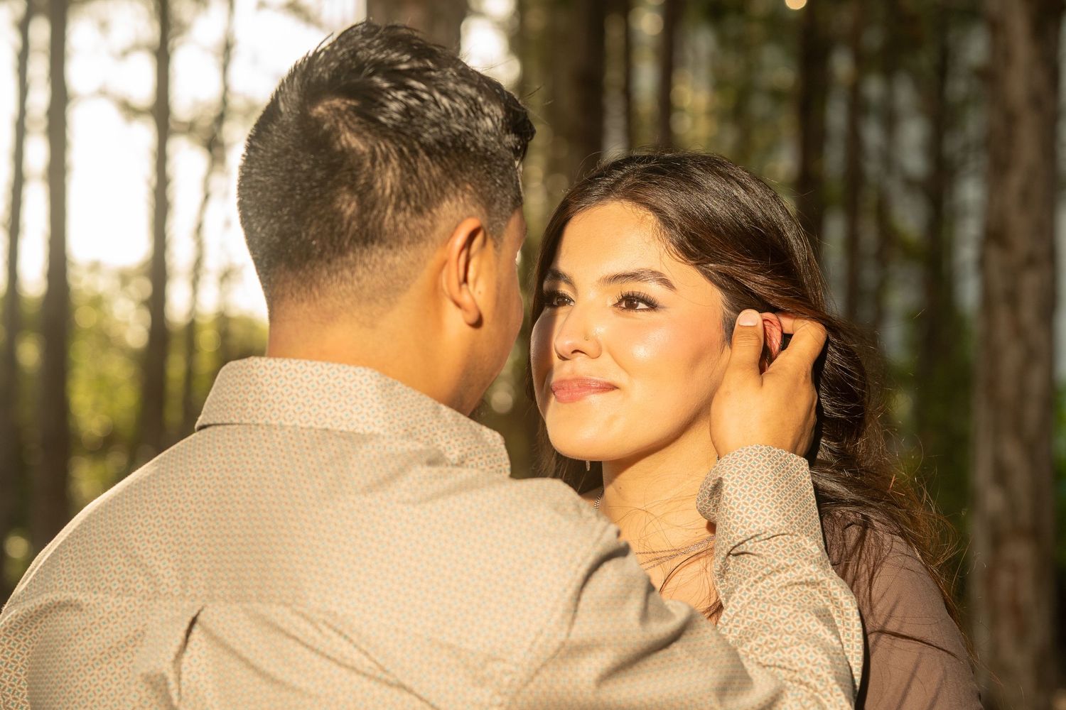 A couple shares a romantic moment in a sunlit forest, with the man tenderly touching the woman's face.