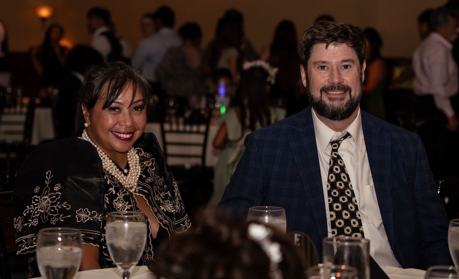 Two elegantly dressed guests smile at a formal banquet dinner event, seated at a decorated table.
